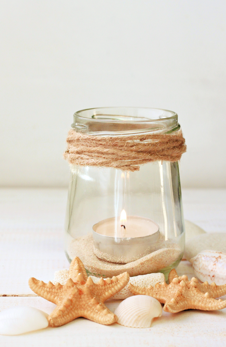 coastal home decor-beach inspired decor. A clear glass jar with just string wrapped around the top with sand inside at the bottom and votive candle. Seashells and shells are scattered around.